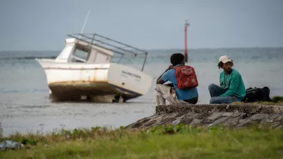 Two young fishers sit by the sea in front of their boat on the sand.